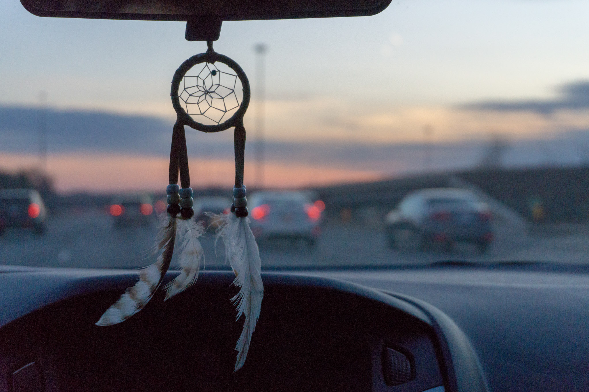 Dreamcatcher with feathers hanging from a car rearview mirror with a blurred sunset sky and highway traffic in the background.