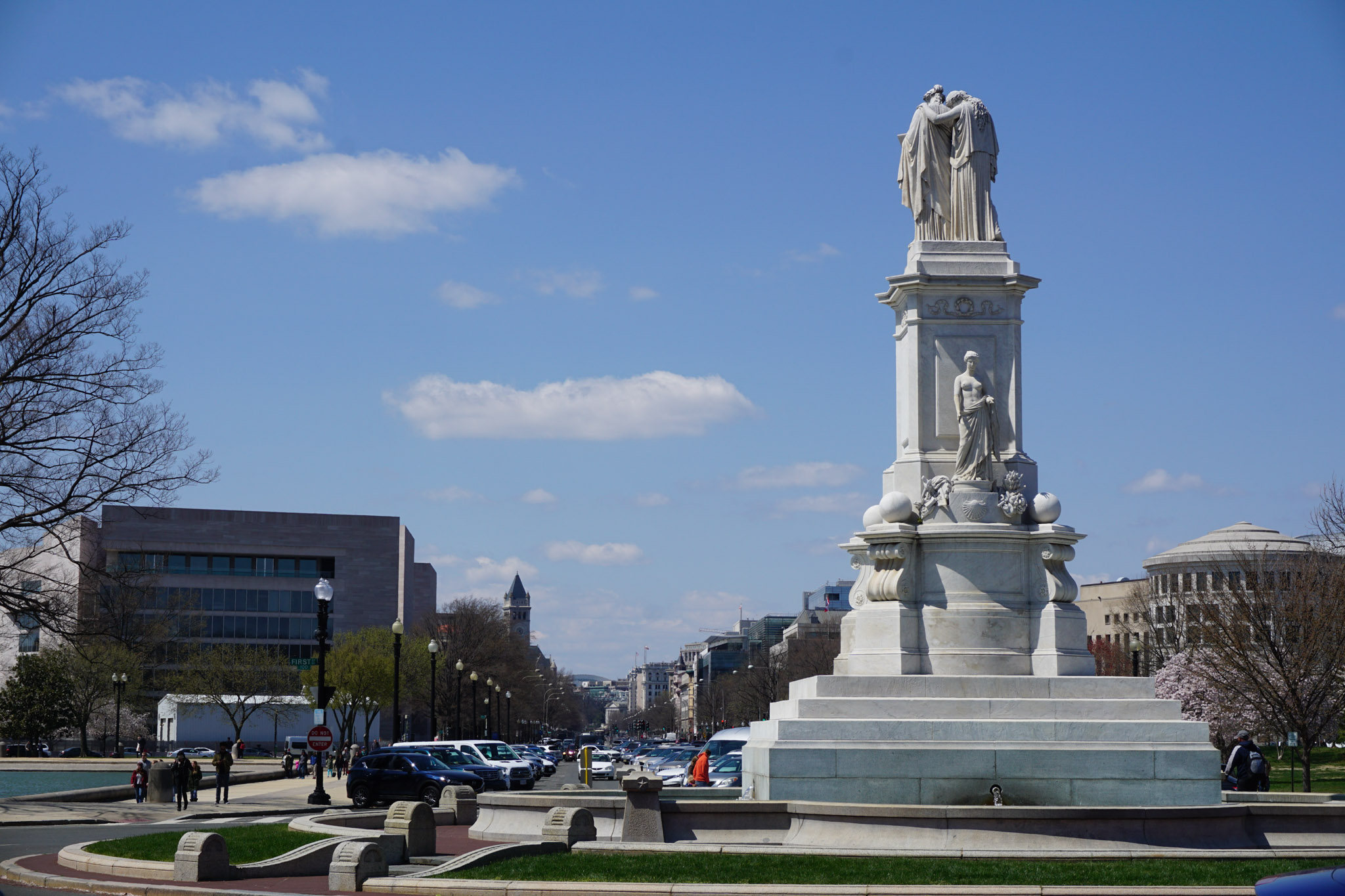 A white marble monument near the U.S. Capitol in Washington, DC, standing above a traffic circle under a bright blue sky with scattered clouds.