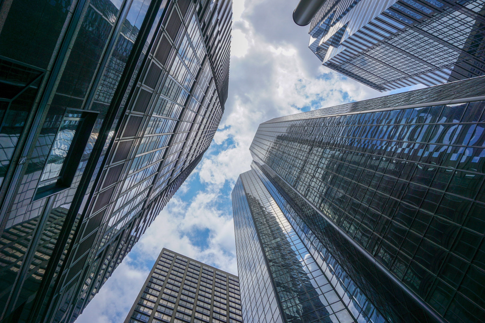 Looking up at modern skyscrapers in downtown Philadelphia, with reflective glass facades framing a patch of blue sky and drifting clouds.