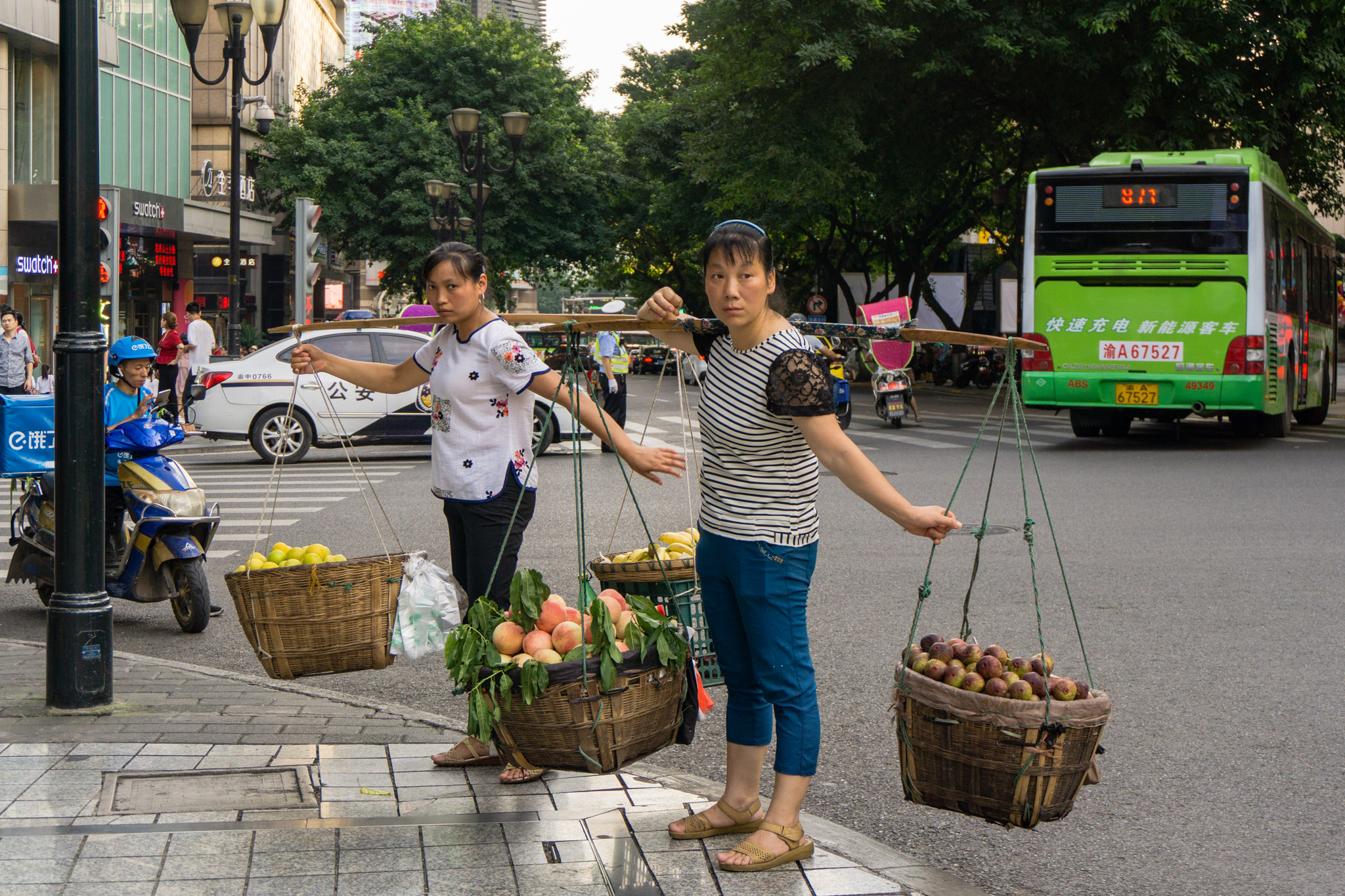 Chongqing street vendors carrying fruit baskets with shoulder poles on a busy city corner, with buses, scooters, and pedestrians in the background.
