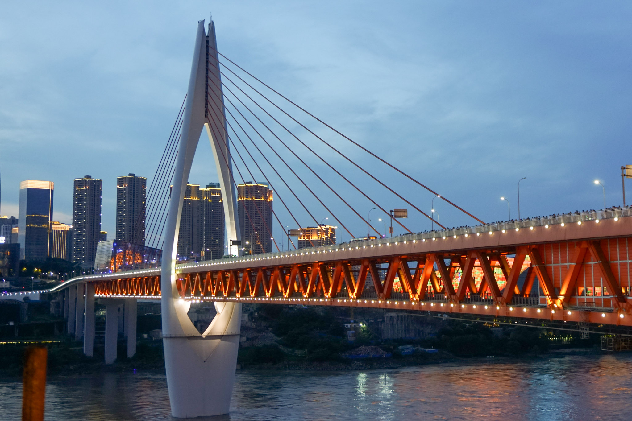 Qiansimen Bridge over the Jialing River in Chongqing at blue hour, with orange-lit trusses, a white cable-stayed tower, and city high-rises in the background.