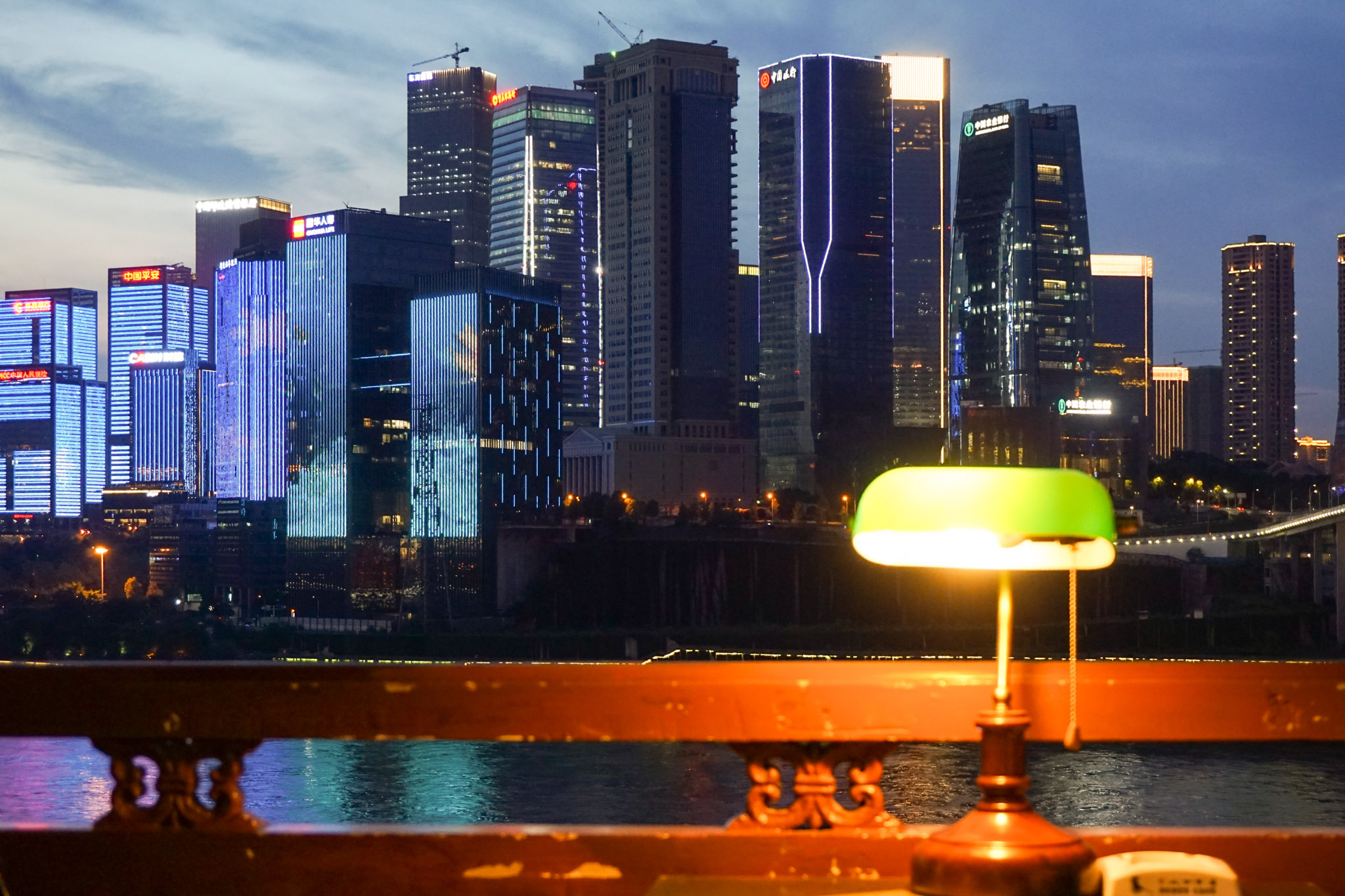 Chongqing skyline at dusk with illuminated skyscrapers across the river, viewed from a terrace with a glowing green desk lamp in the foreground.