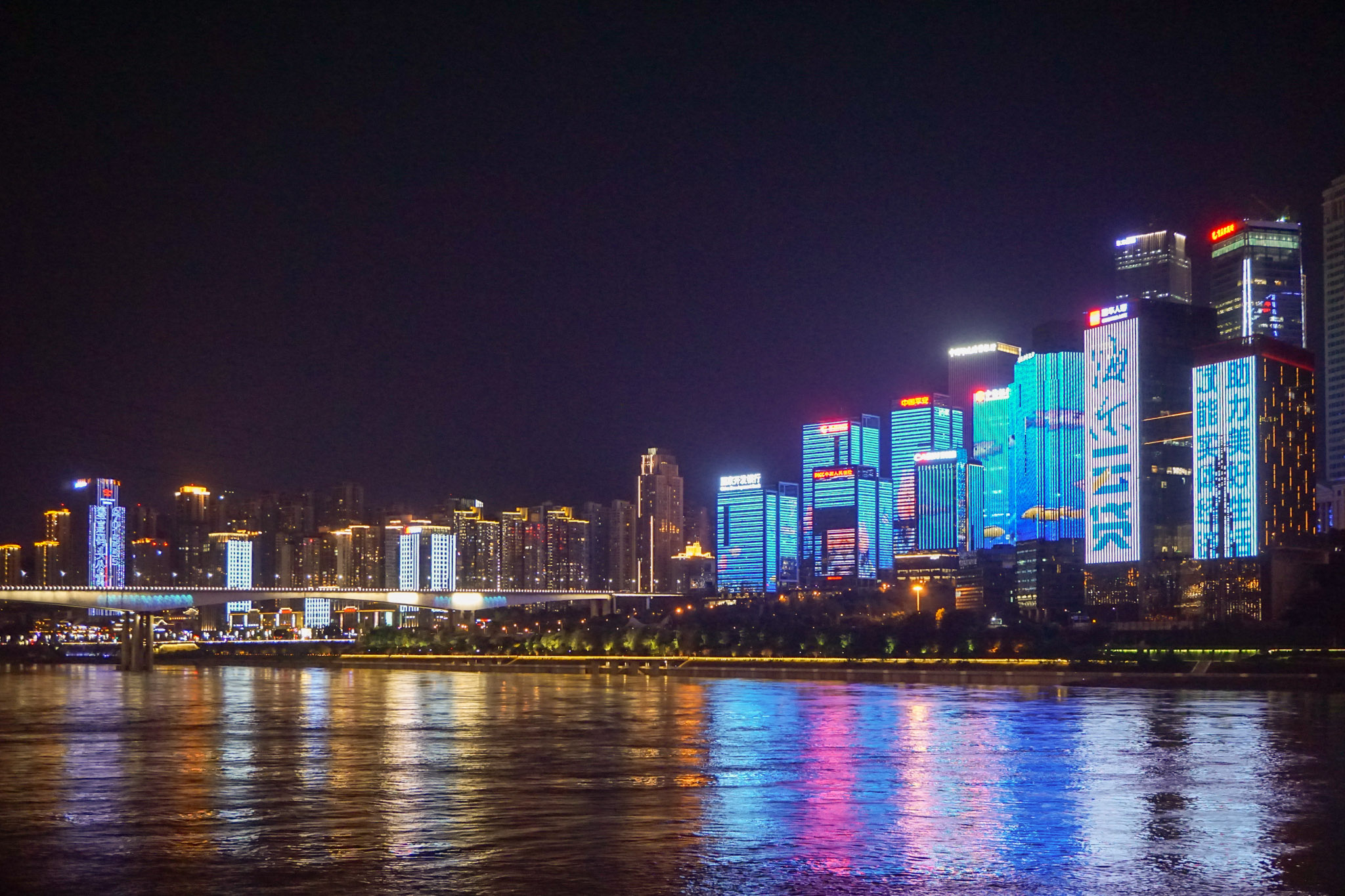 Chongqing riverside skyline at night, with brightly lit skyscrapers and colorful neon reflections shimmering on the river.
