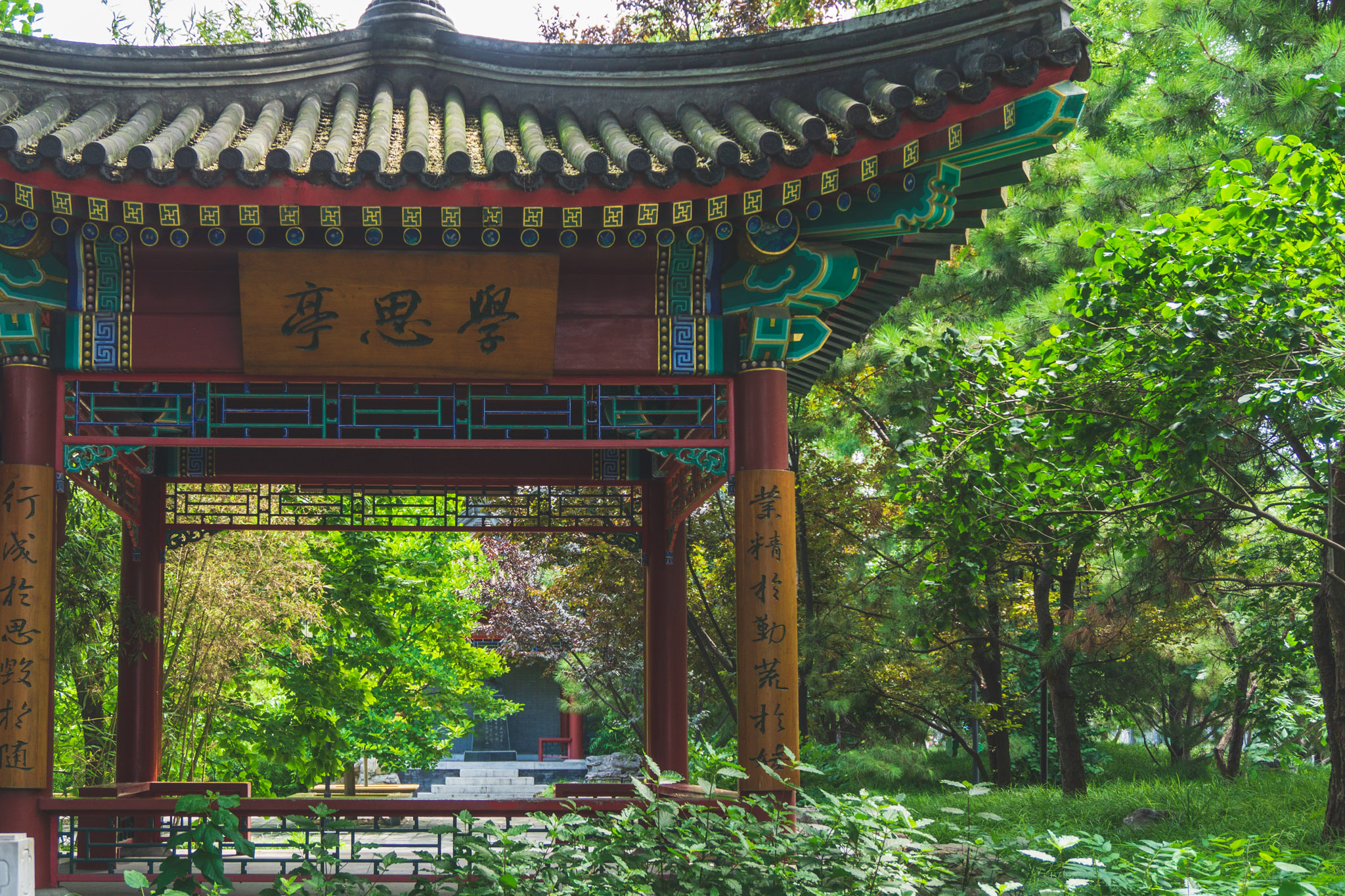 Traditional Chinese pavilion in Beijing surrounded by lush summer greenery, with ornate painted beams and a curved tiled roof.